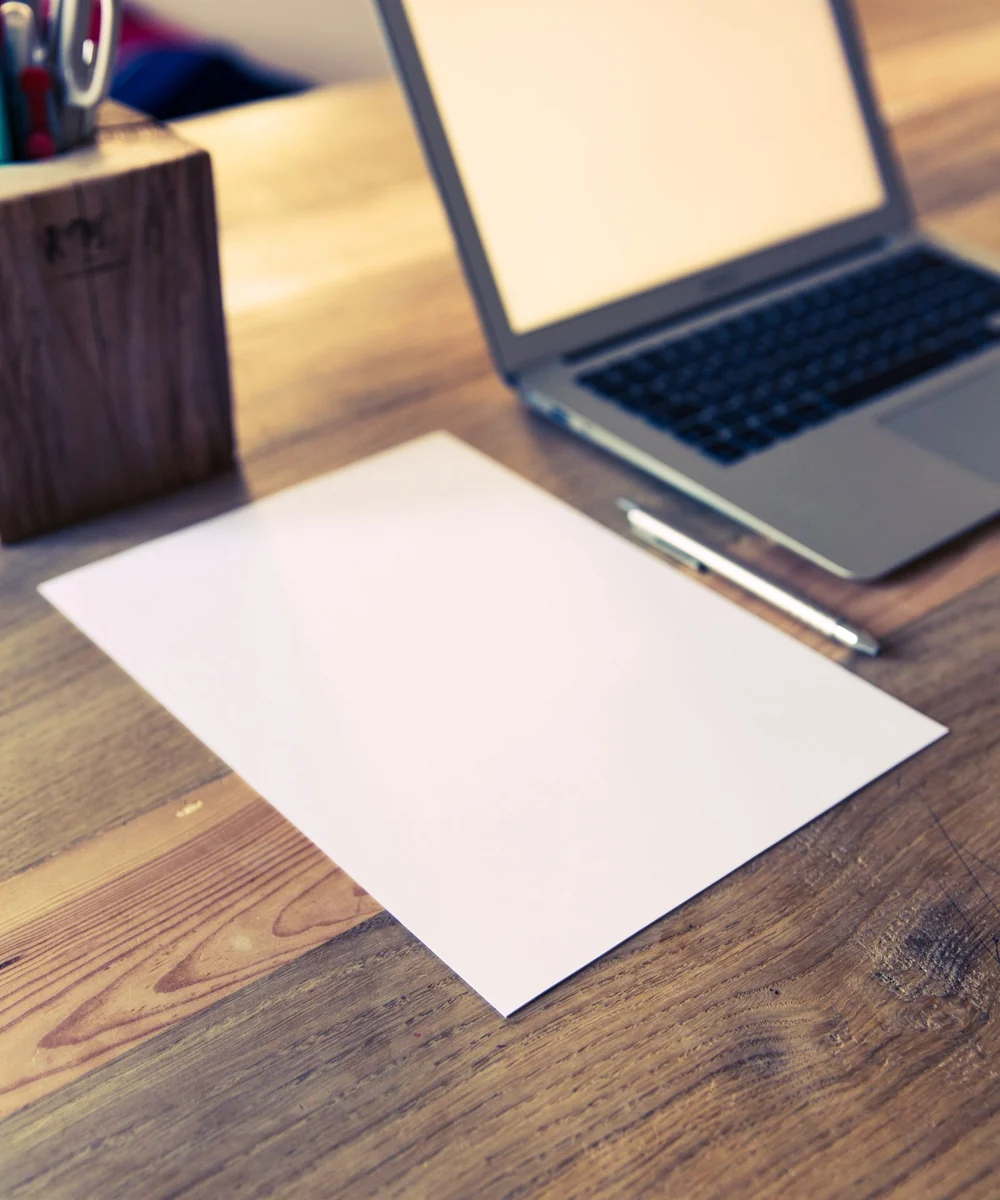 Papel em branco, caneta e notebook sobre uma mesa de madeira, sem pessoas.